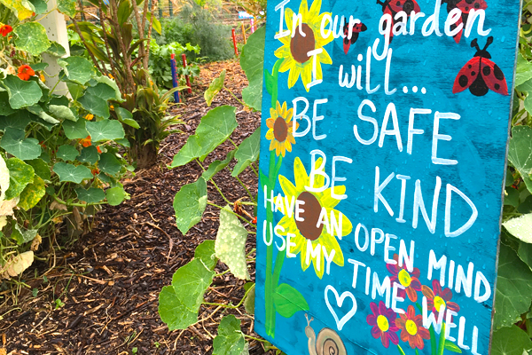 In a garden, a blue handpainted depicts sunflowers and ladybugs. Its text reads: In our garden I will be safe, be kind, have an open mind, use my time well.
